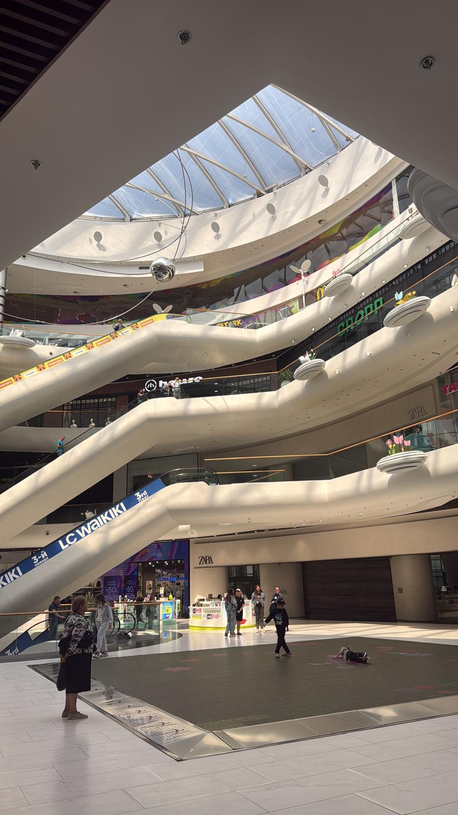 Foto der Rolltreppen im Inneren der fünfstöckigen Mall. Das Dach ist eine Glaskuppel und im Erdgeschoss können Kinder auf einem Monitor auf dem Boden Spiele spielen.