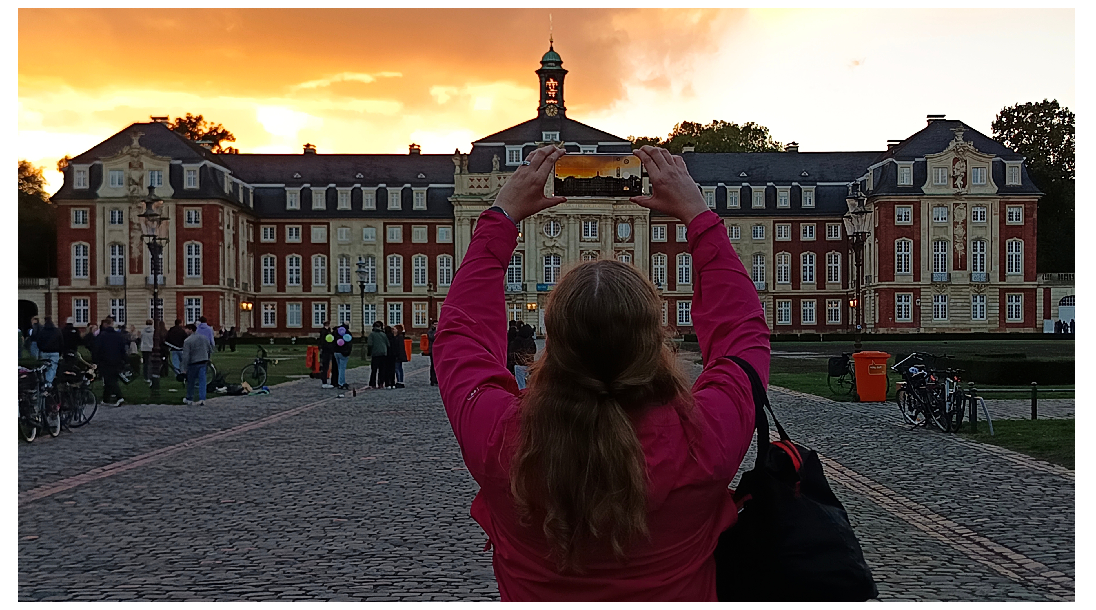 Menschen machen Fotos vom Schloss. Hinter dem Schloss in Münster geht die Sonne unter und die Wolken leuchten in einem dunklen orangenen/goldenen Licht. Vor dem Schloss steht eine Frau mit dem Rücken zur Kamera, die ein Handy hoch hält und ebenfalls ein Foto vom Schloss schießt.