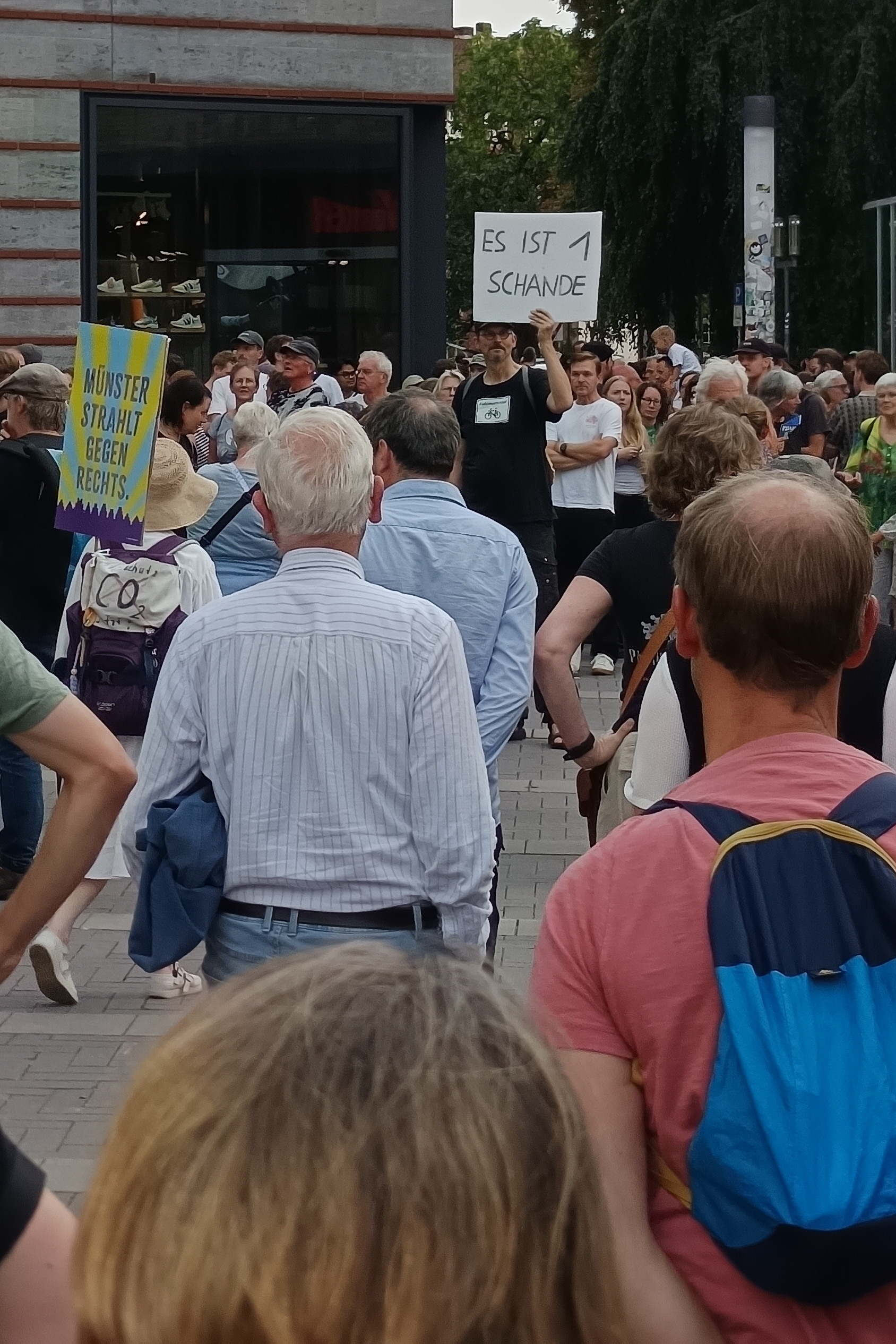 Foto aus der Menschenmenge. Plakate mit Aufschriften wie 'Münster strahlt gegen rechts'. Ein großer Mann hält ein Plakat mit dem Text 'Es ist 1 Schande' hoch.