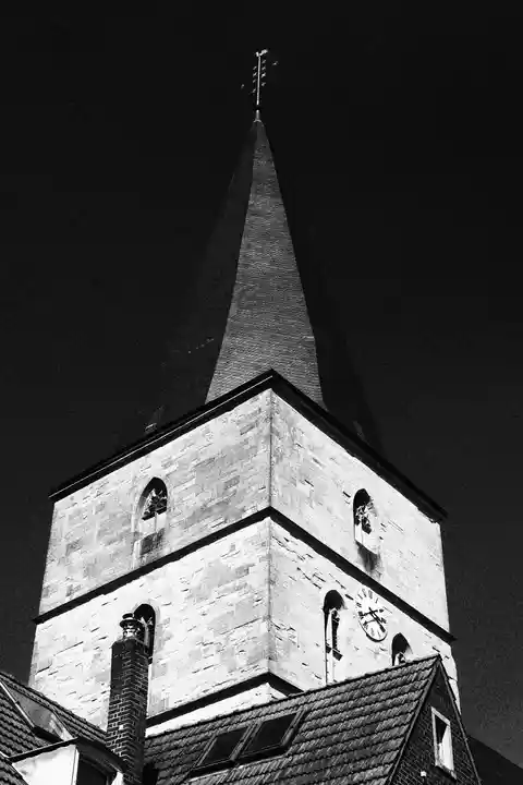 Schwarz-Weiß Foto eines Kirchturmes aus der Perspektive direkt davor, wodurch der Turm mächtig wirkt. Der Himmel ist schwarz und das dunkle Dach der Kirche kaum zu erkennen. Dafür strahlt das helle Mauerwerk um so mehr.