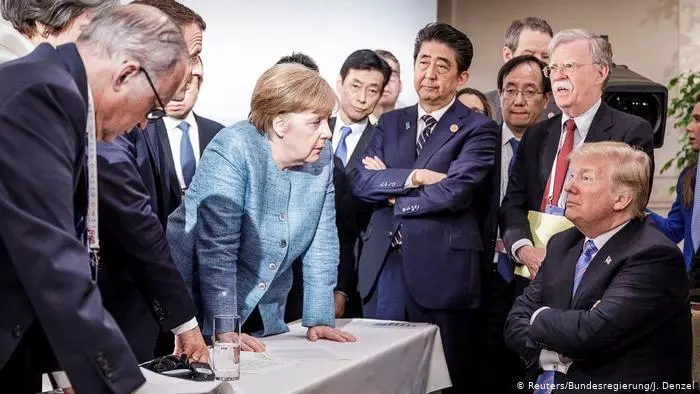 Foto vom G7-Gipfel auf dem Trump mit verschränkten Armen trotzig an einem Tisch sitzt und sich Angela Merkel von der anderen Seite rüber beugt