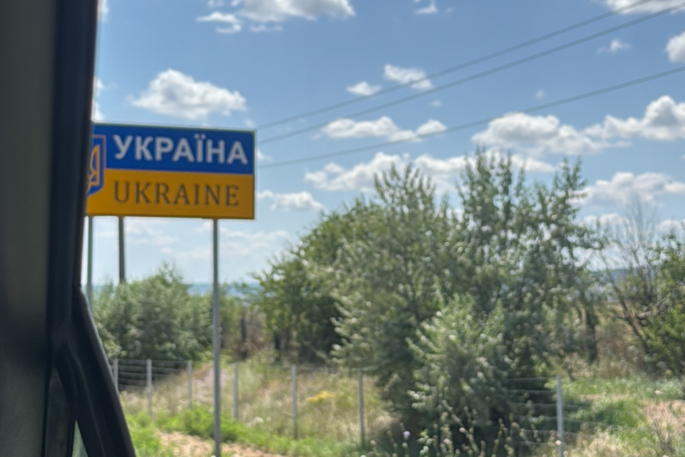 Foto aus dem Beifahrer-Fenster der sonnigen Landschaft mit Büschen und einem Blau-Gelben Schild, welches das Emblem der Ukraine sowie den Namen des Landes auf Lateinischen und Kyrillischen Buchstaben enthält.